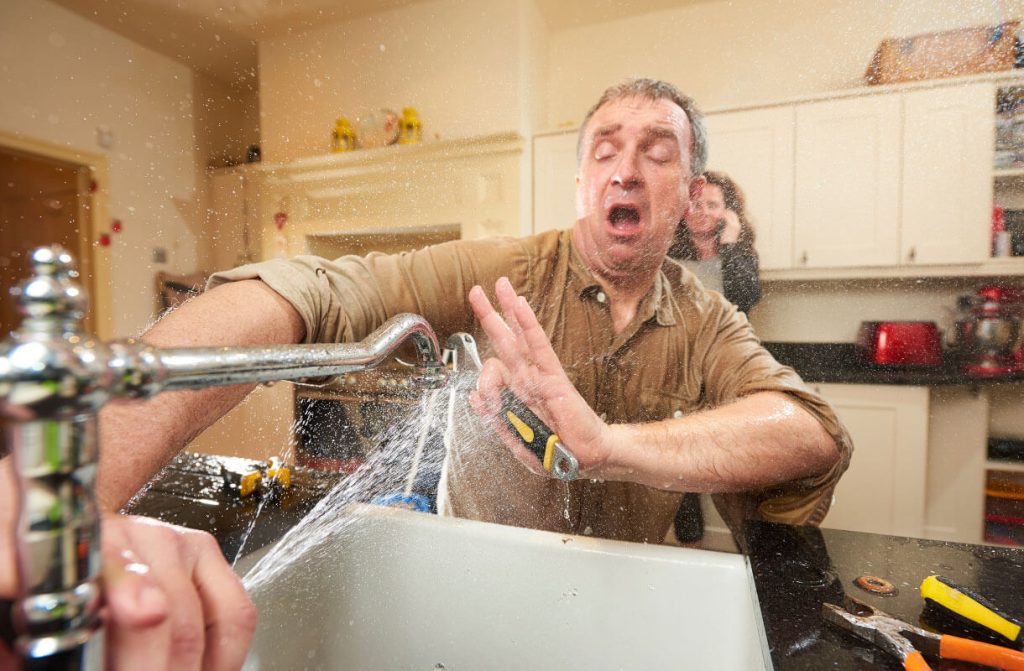 Man reacting to burst kitchen faucet spraying water during plumbing emergency